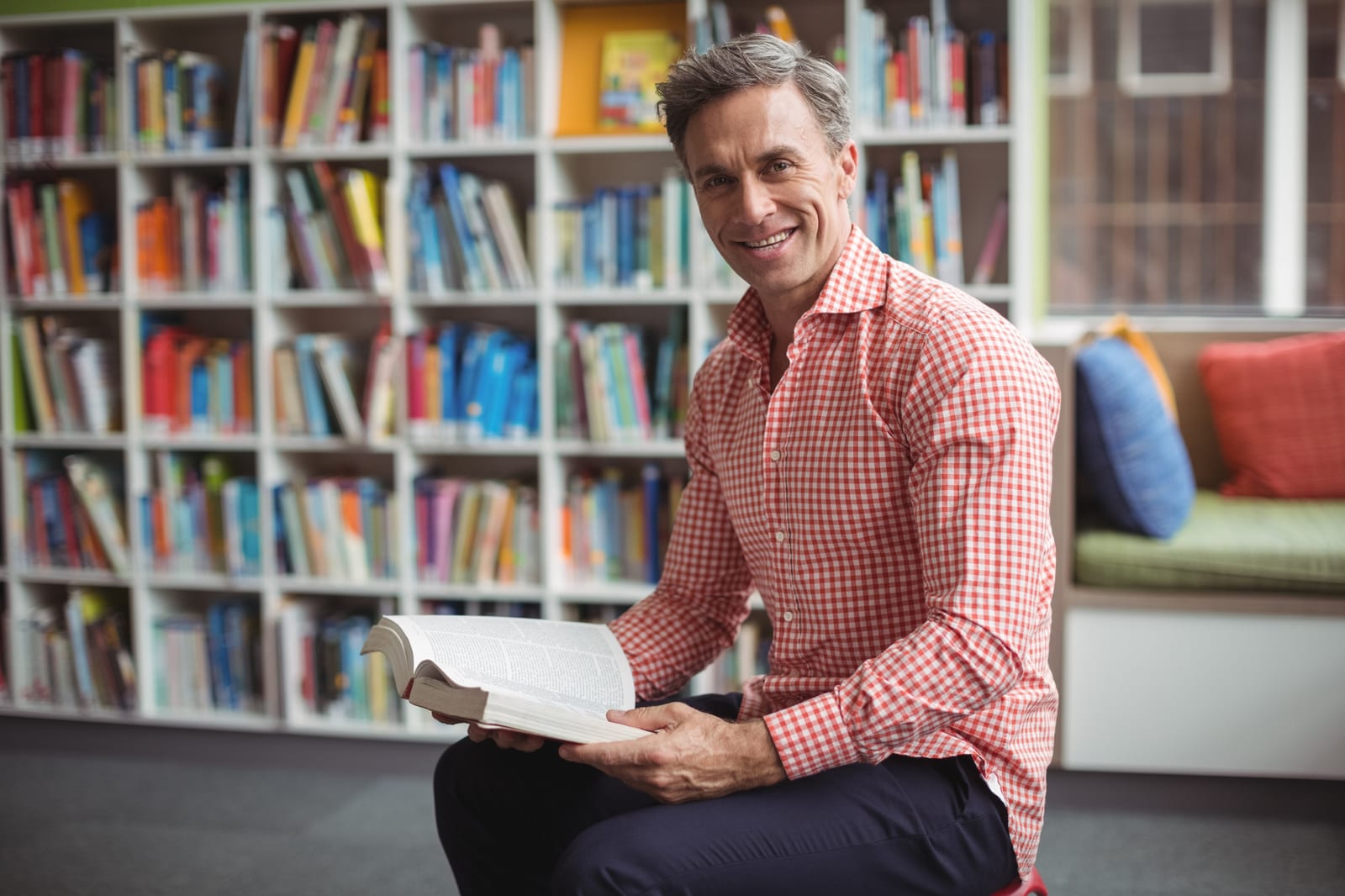 Homem sorridente segurando um livro aberto, sentado em frente a estantes coloridas de biblioteca